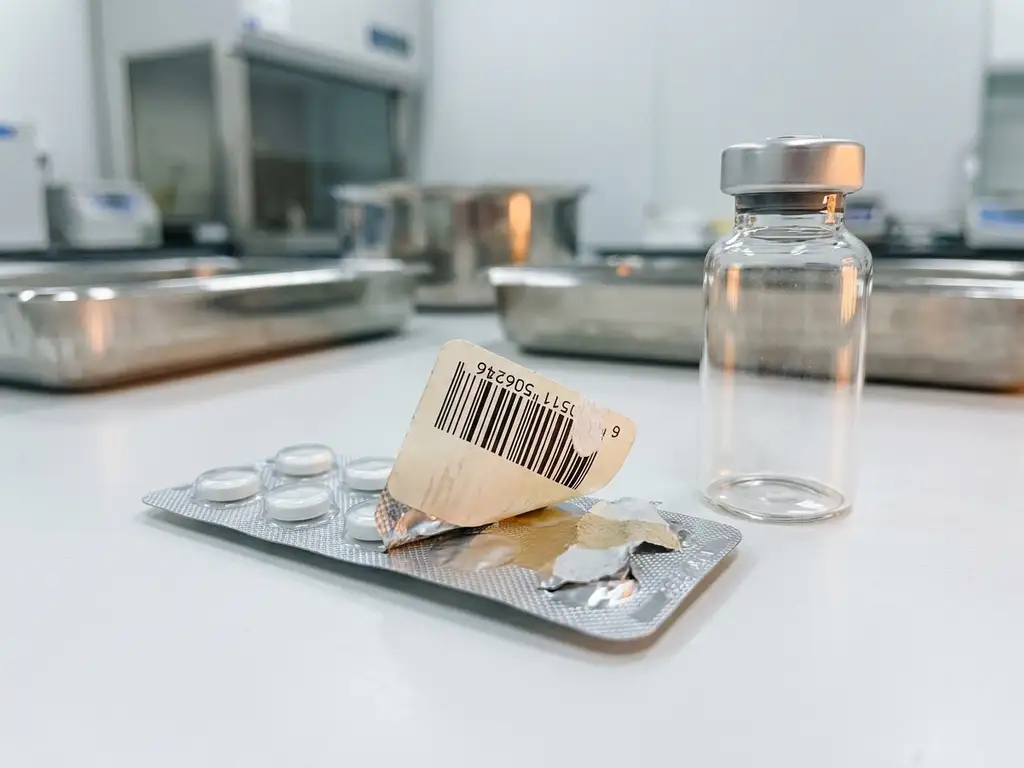 Pharmazeutische Blisterpackung und Glasampulle auf weißer Laborfläche mit abgelöstem Barcode-Etikett, klinische Umgebung.